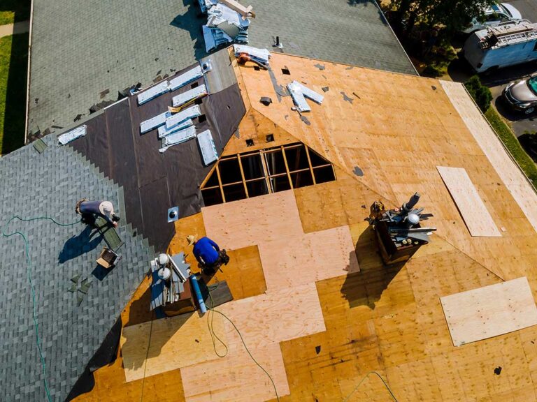 Two workers repairing shingles on a residential roof with tools and safety harnesses.