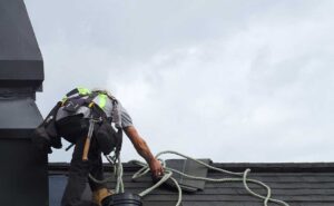 A roofer inspects metal flashing around a plumbing vent on a shingled roof.